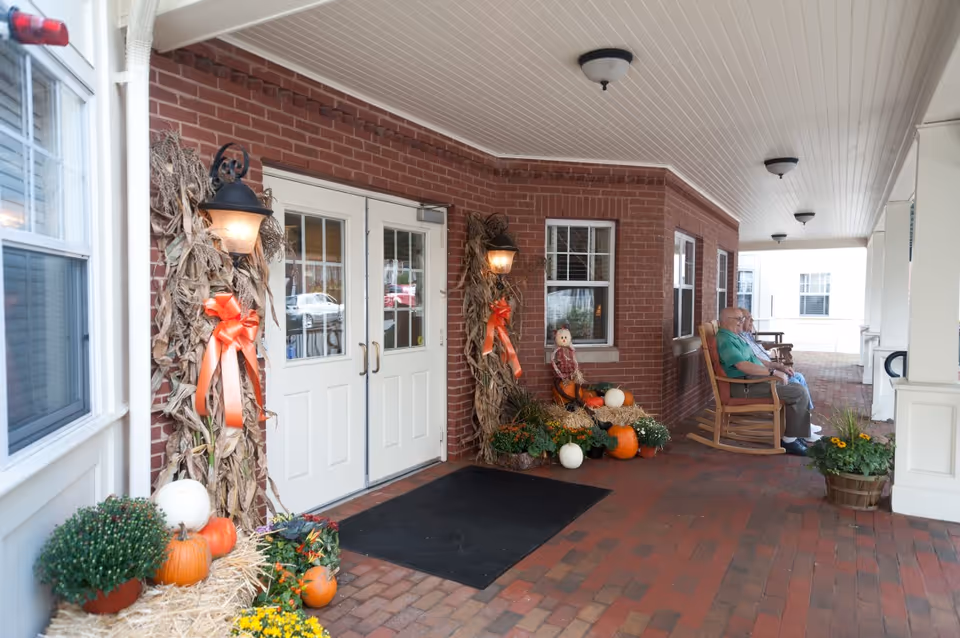 Brick building entrance with a covered porch, fall decorations, pumpkins, and a seated resident.