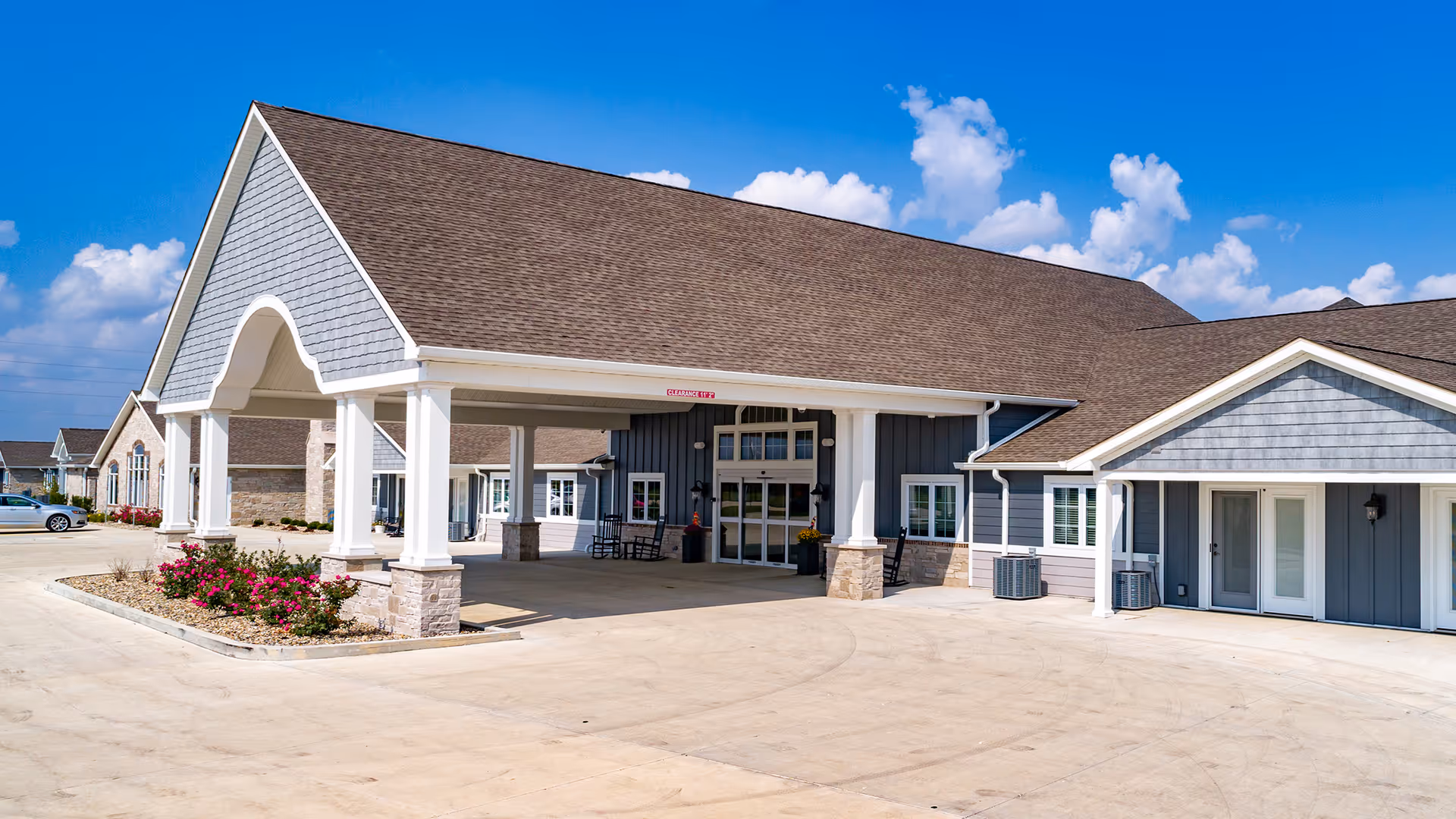Front exterior view of Villas of Holly Brook Assisted Living & Memory Care building in Jerseyville, IL, showing a covered entrance with white pillars, a landscaped area with flowers, and a clear blue sky with some clouds.