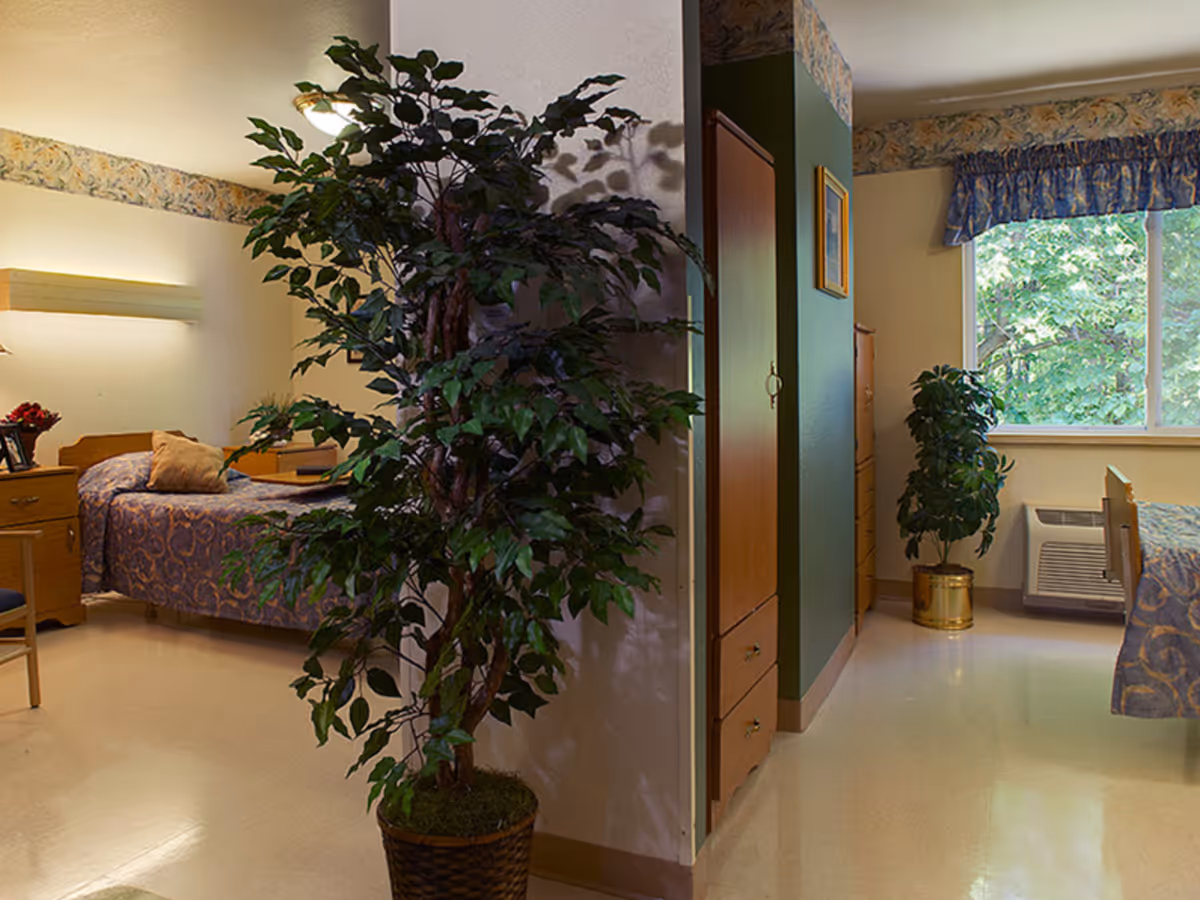 Interior view of a senior living facility bedroom with two beds, each covered with patterned bedspreads. There are two large potted plants, one in the foreground and one near a window with blue curtains. The room has light-colored walls with a floral border near the ceiling, wooden furniture including dressers and nightstands, and a heating or air conditioning unit below the window.