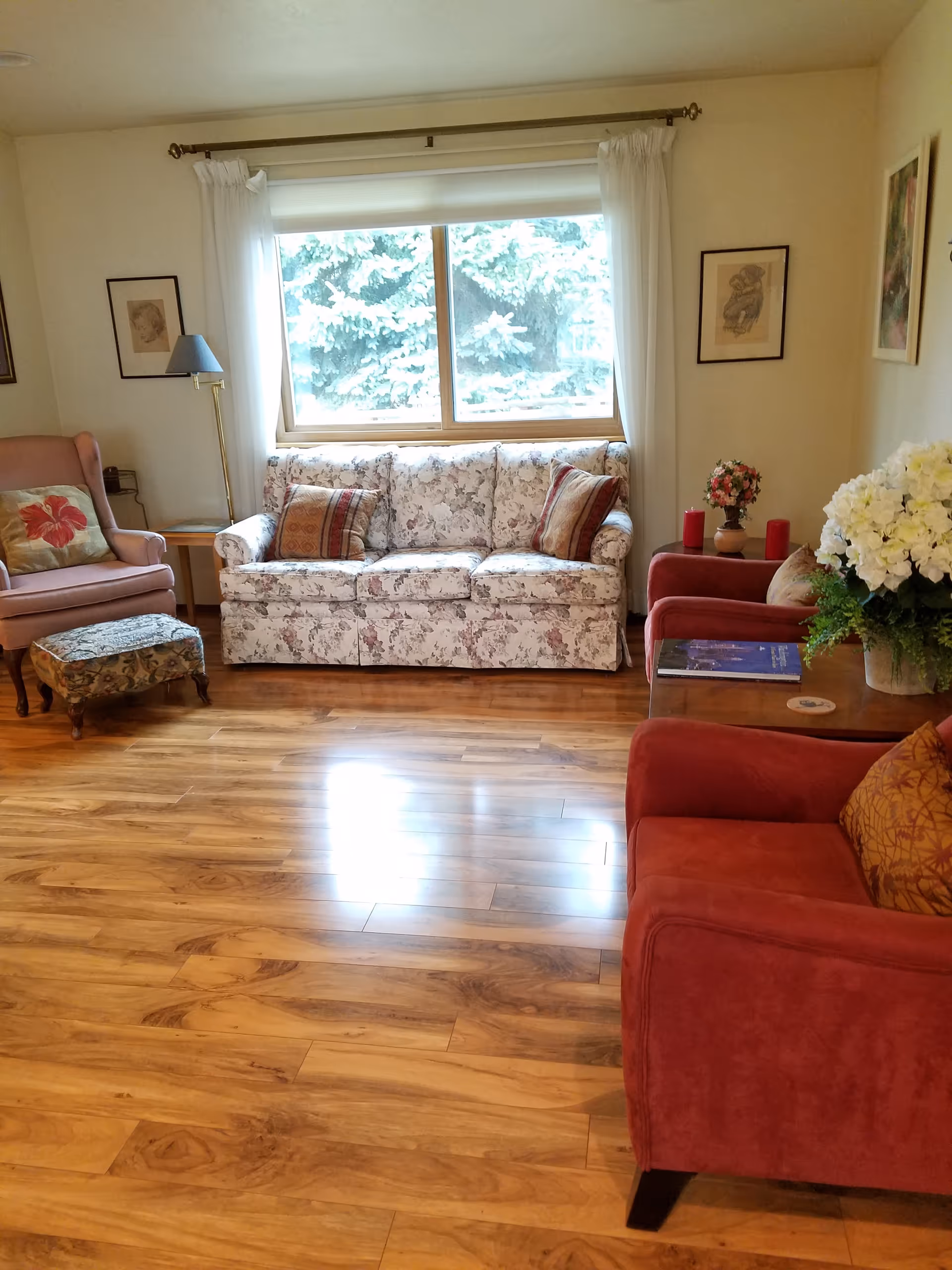 Cozy living room with floral sofa, red chairs, large window, and wood floors.