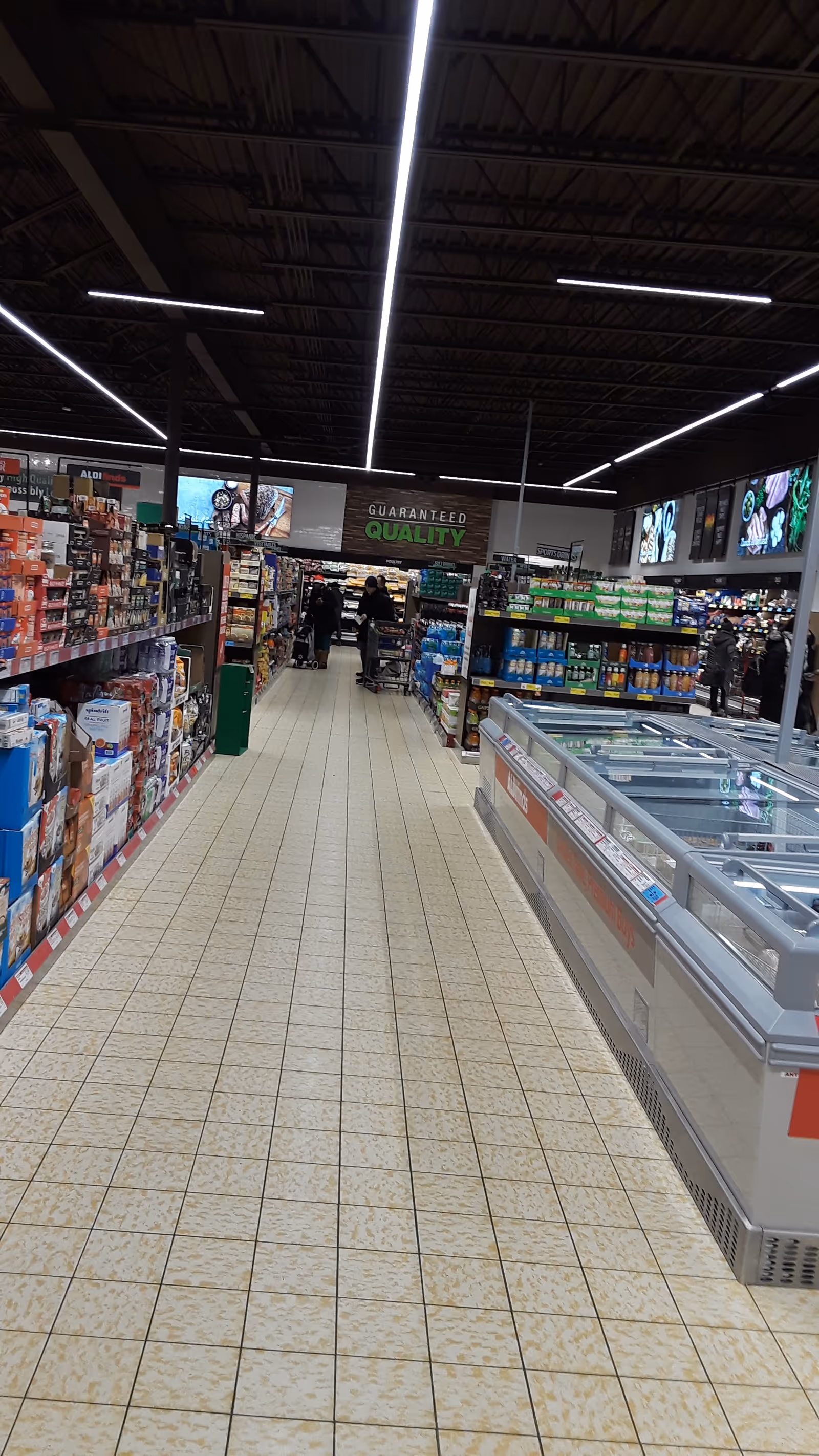 A long supermarket aisle with shelves, a freezer section, and bright overhead lights.