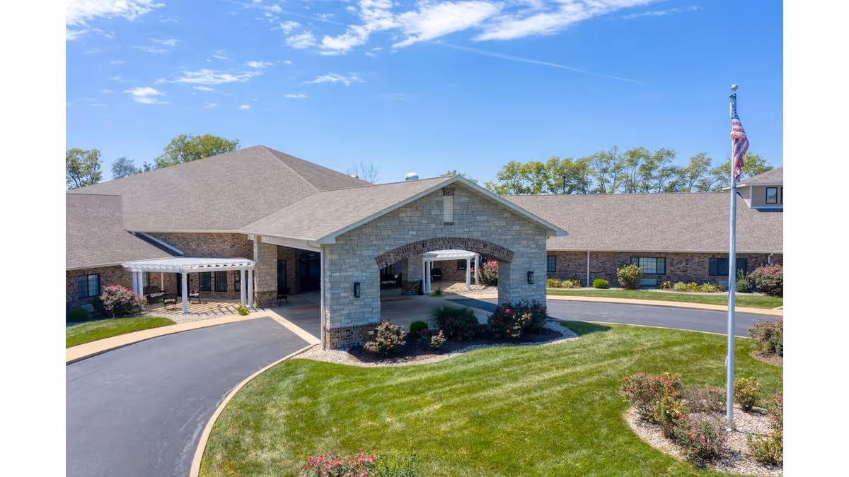 Exterior view of Cedarhurst of Breese senior living facility showing a large building with a covered entrance, well-maintained green lawn, flower beds, and an American flag on a flagpole under a clear blue sky.