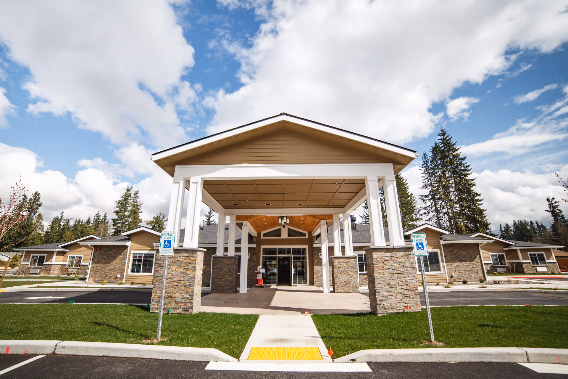 Front entrance of a single-story memory care building with a covered porte-cochère, stone columns, accessible parking signs, and landscaped grounds under a partly cloudy sky.