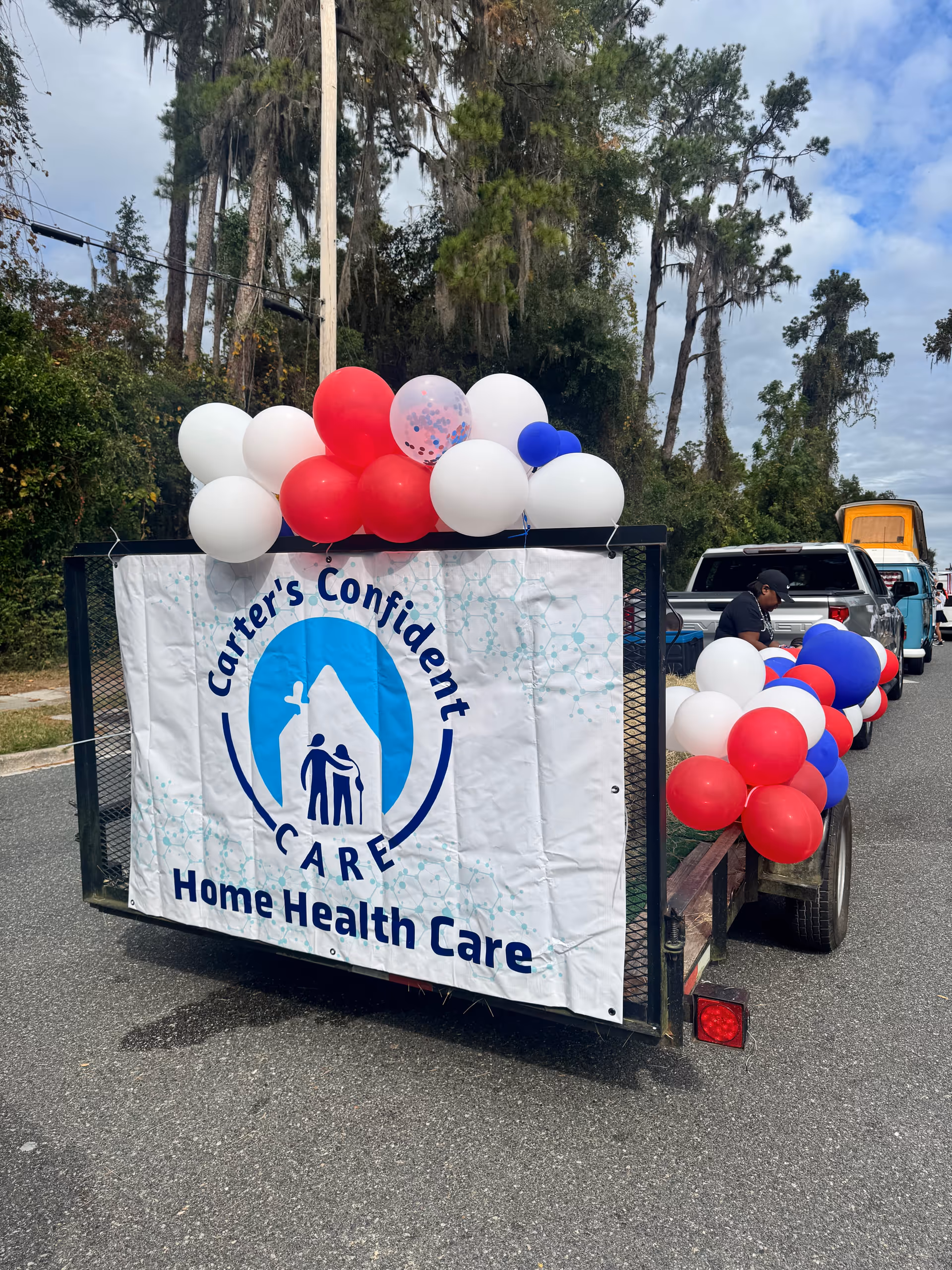 A trailer with Carter's Confident Care Home Health Care banner and red, white, blue balloons outdoors.