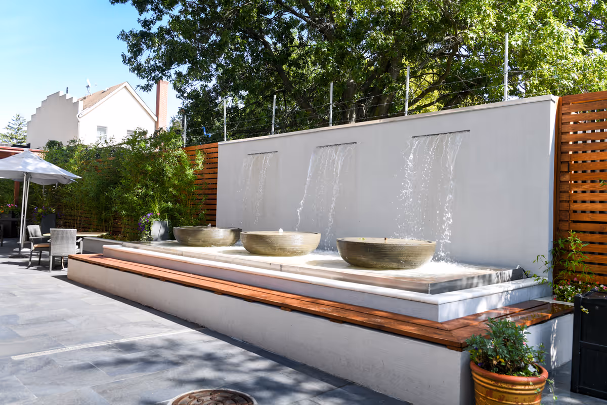 Outdoor courtyard area with a modern water feature consisting of three streams of water flowing into large round basins. There are wooden benches along the water feature, potted plants, and patio furniture with an umbrella. Trees and a wooden fence provide a natural and private setting.