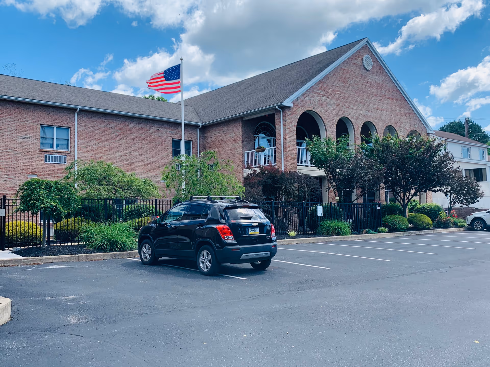 Exterior view of a two-story brick building with arched windows and a balcony, surrounded by trees and shrubs. An American flag is flying on a flagpole in front of the building. A black SUV is parked in the parking lot in front of the building under a partly cloudy blue sky.