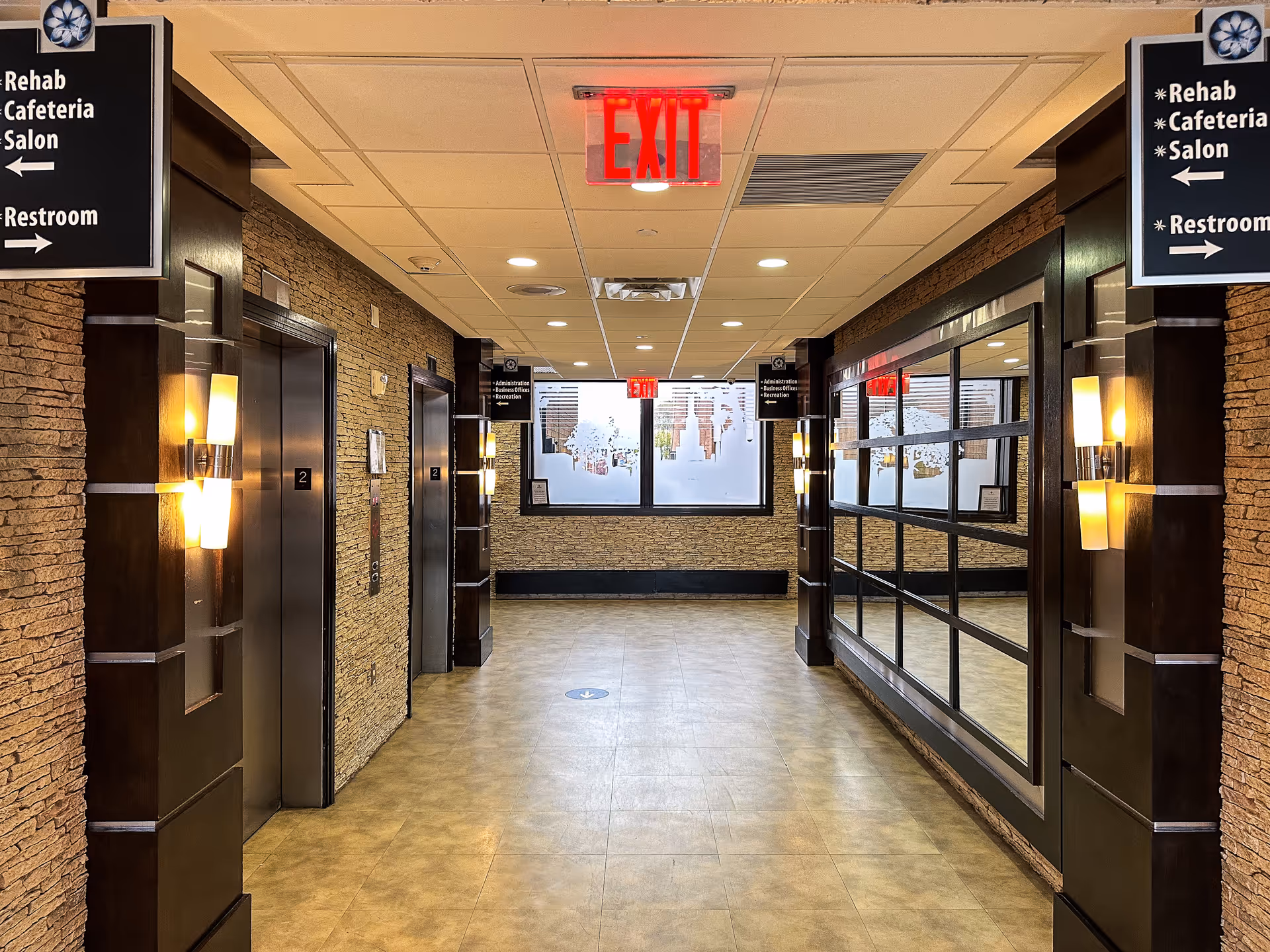 Interior senior-facility hallway with elevators, wall lights, and an illuminated EXIT sign.