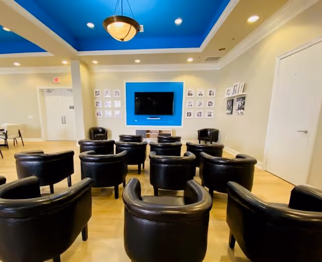 A communal lounge with rows of black leather armchairs facing a wall-mounted TV set in a blue-accented wall beneath a coffered blue ceiling.