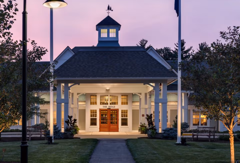 Front entrance of a senior living building at dusk featuring a lit covered portico, double wooden doors, flagpoles, benches, and landscaped lawn.