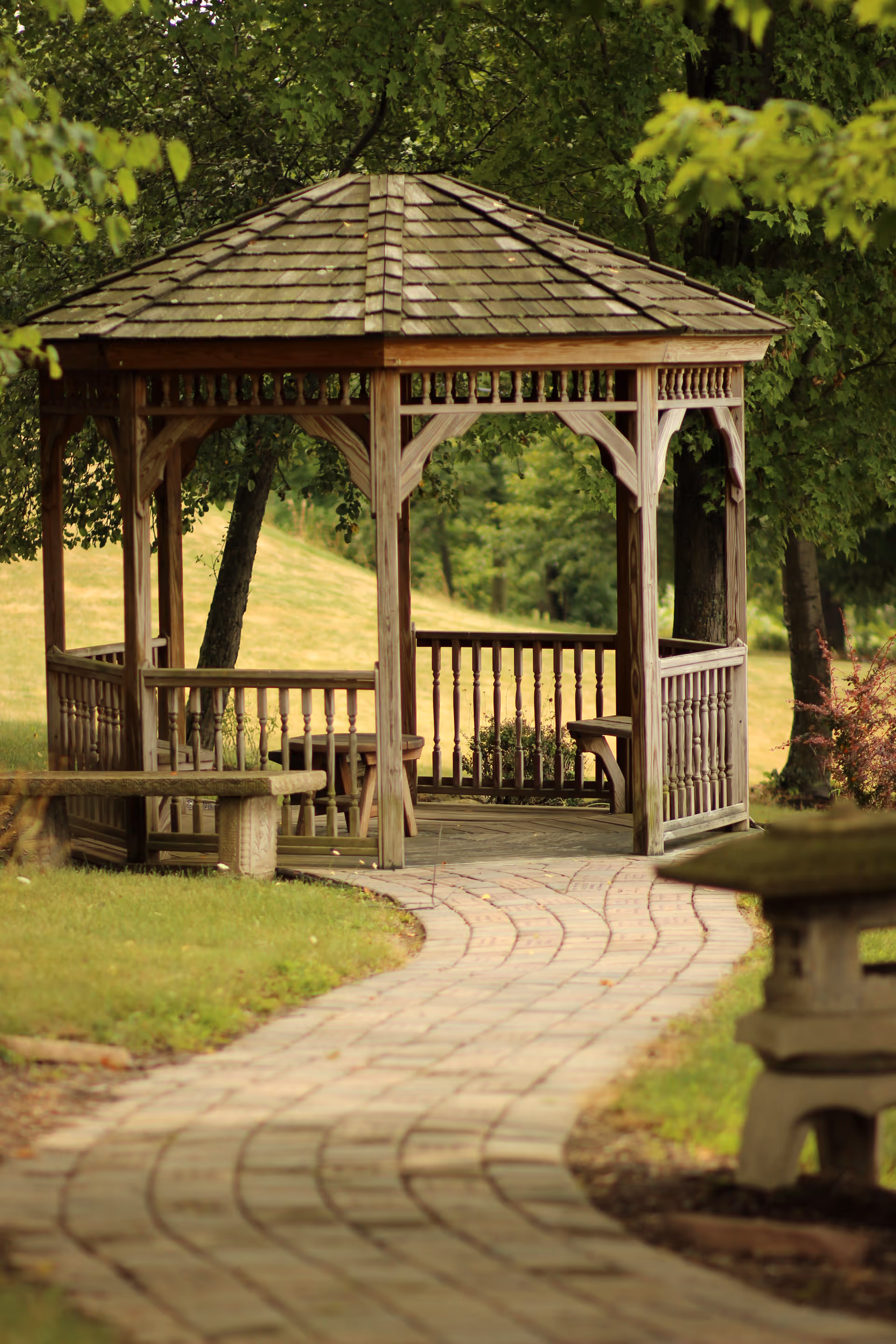 Wooden gazebo beside a curved stone path, surrounded by green trees and grass.