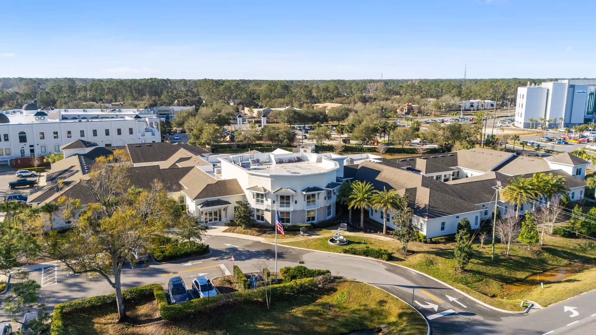 Aerial view of the Artis of Palm Coast senior living facility showing the building's front with a circular driveway, fountain, flagpole, parking and landscaped grounds.