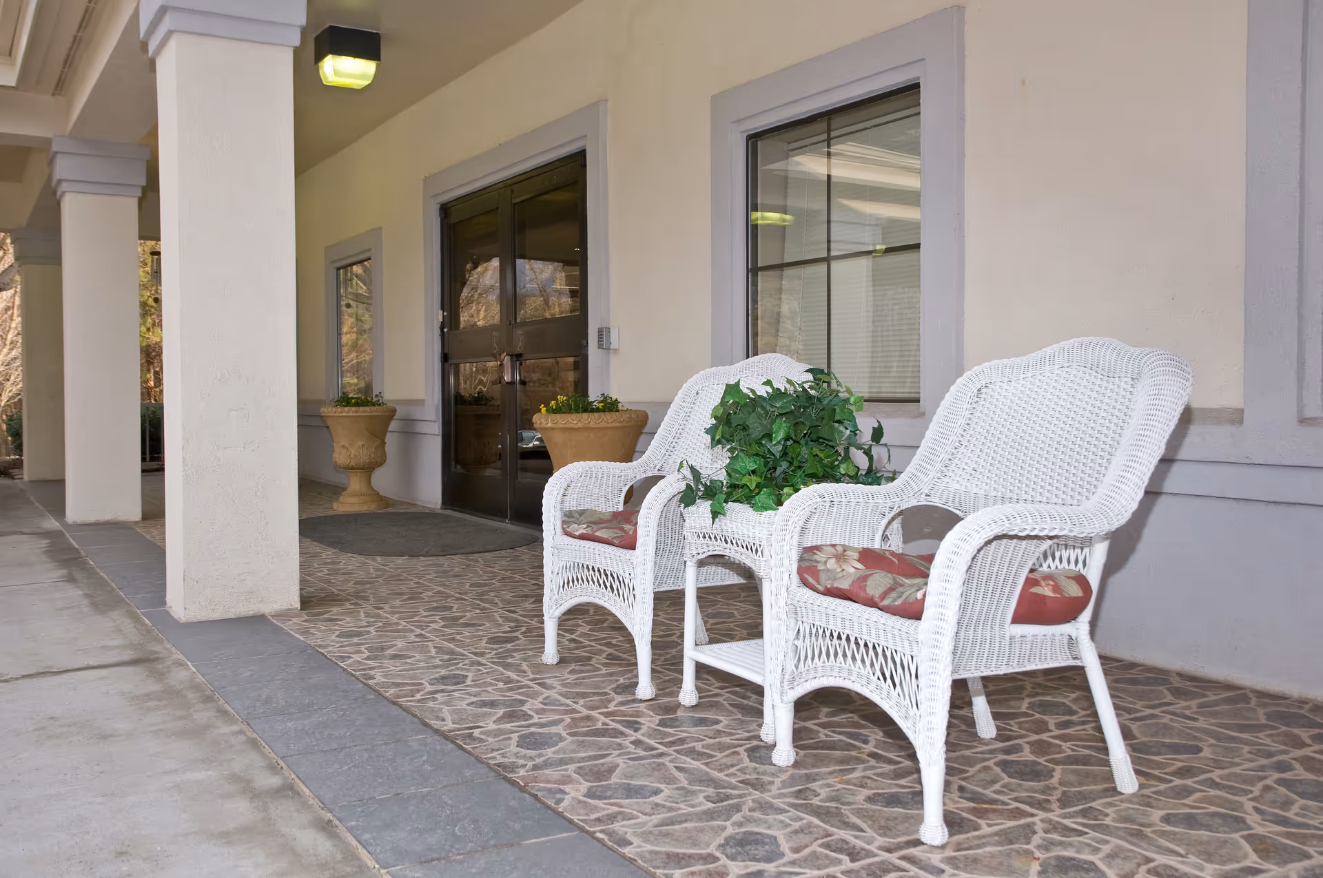 Two white wicker chairs with cushions sit on a stone porch outside the facility entrance.