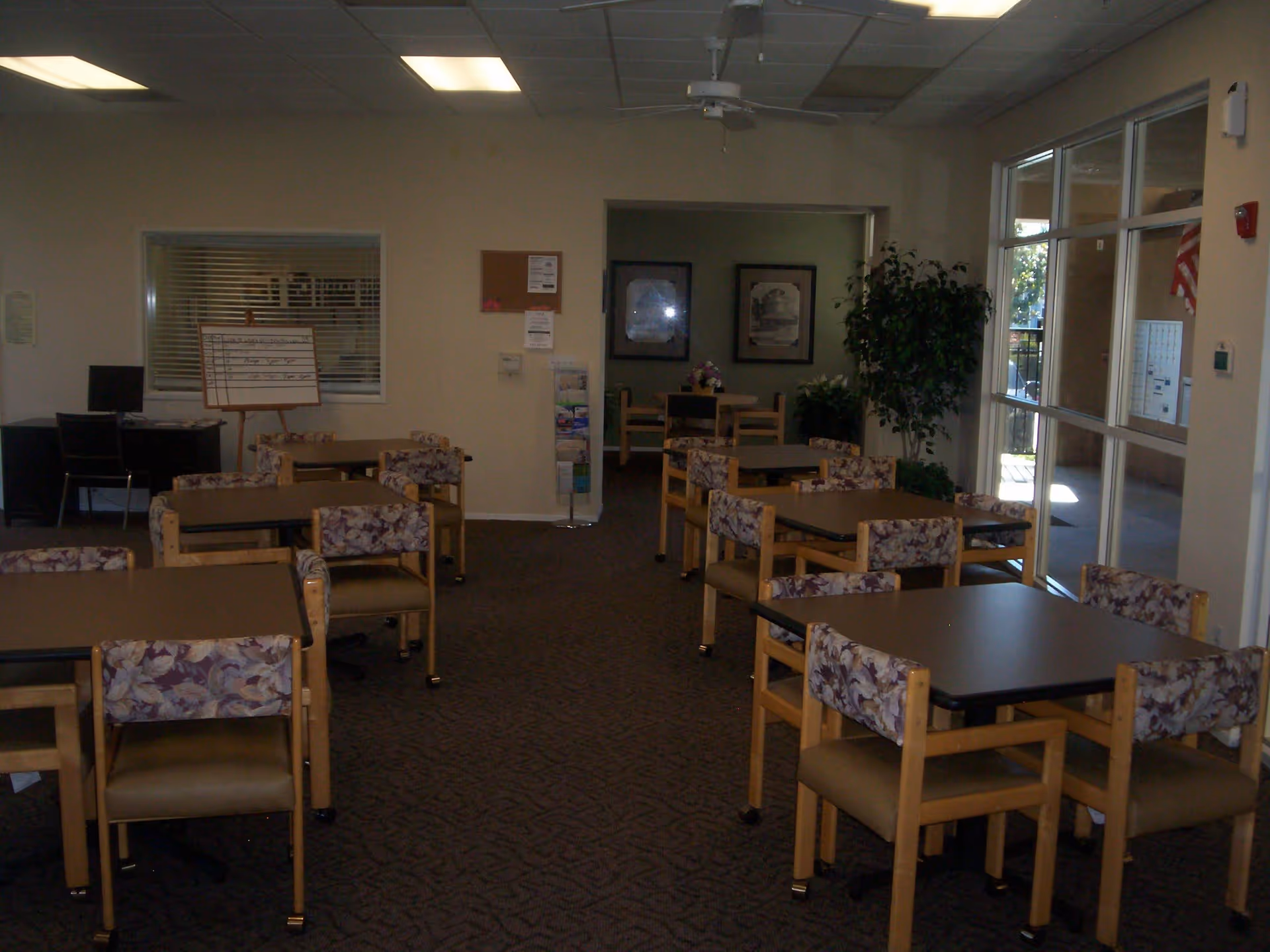 Interior view of a common area in a senior living facility with multiple tables and chairs arranged neatly. The chairs have floral patterned backs and beige seats. There is a ceiling fan, a large window on the right side letting in natural light, and some framed pictures and plants in the background.