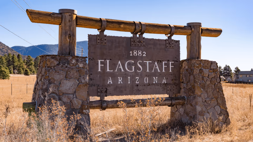 A large rustic wooden and stone sign in a dry grassy field with mountains and trees in the background. The sign reads '1882 FLAGSTAFF ARIZONA'.