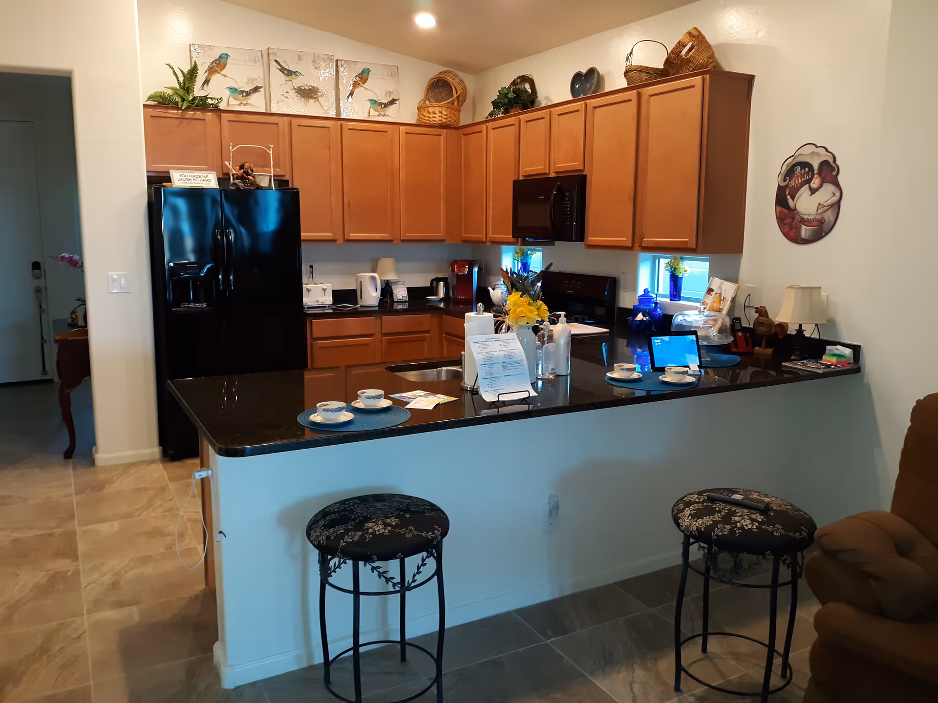 A kitchen area with wooden cabinets, a black refrigerator, black microwave, and black countertop with two stools. The countertop has cups, a tablet, hand sanitizer, and a small flower arrangement. Decorative items and artwork are displayed on top of the cabinets and on the walls.