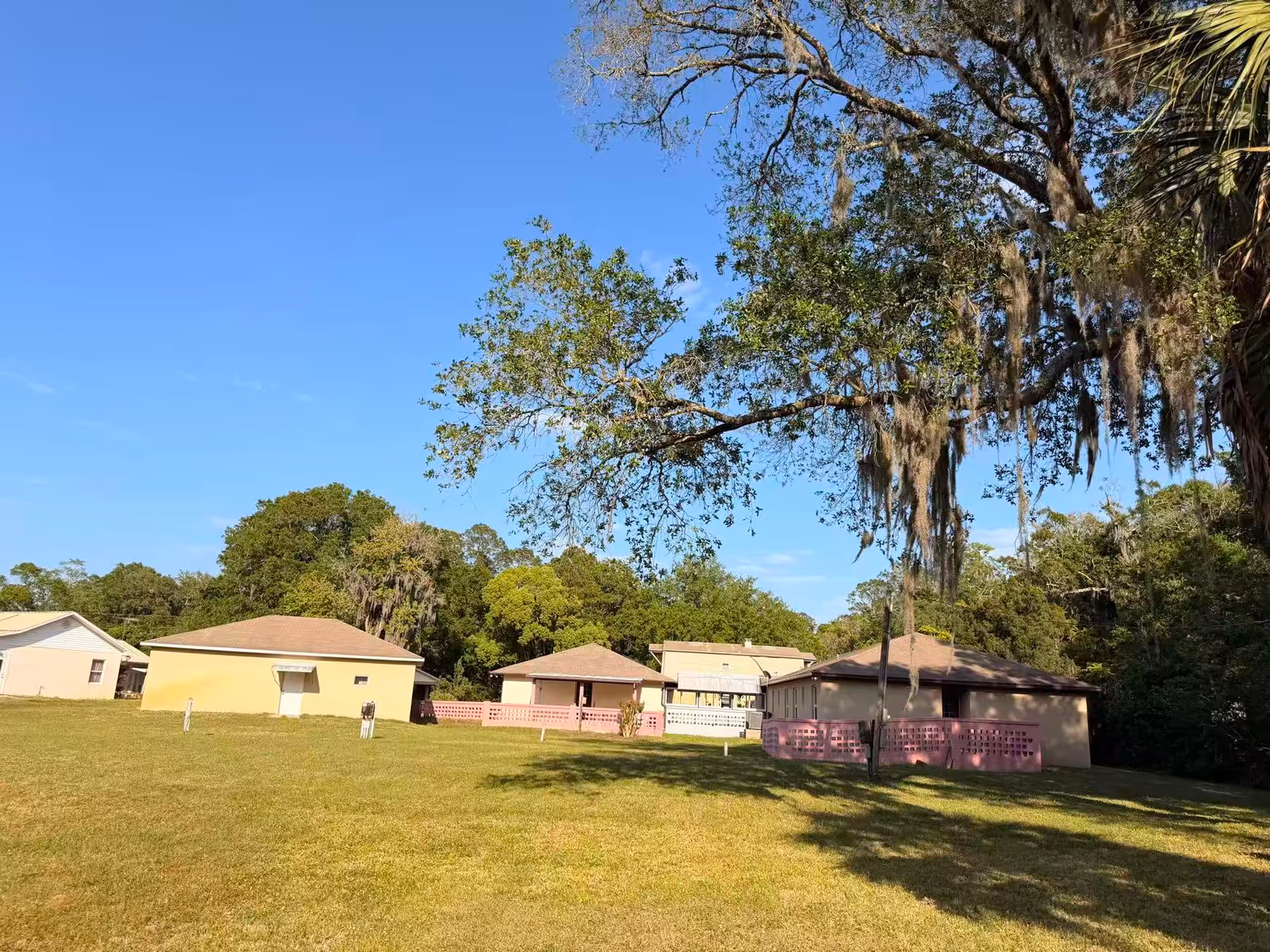 Open grassy yard with pastel single-story buildings, pink fencing, and large trees.
