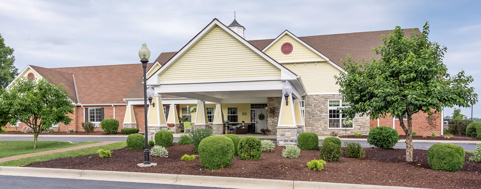 Single-story senior living facility with yellow siding, stone accents, and landscaped entrance.