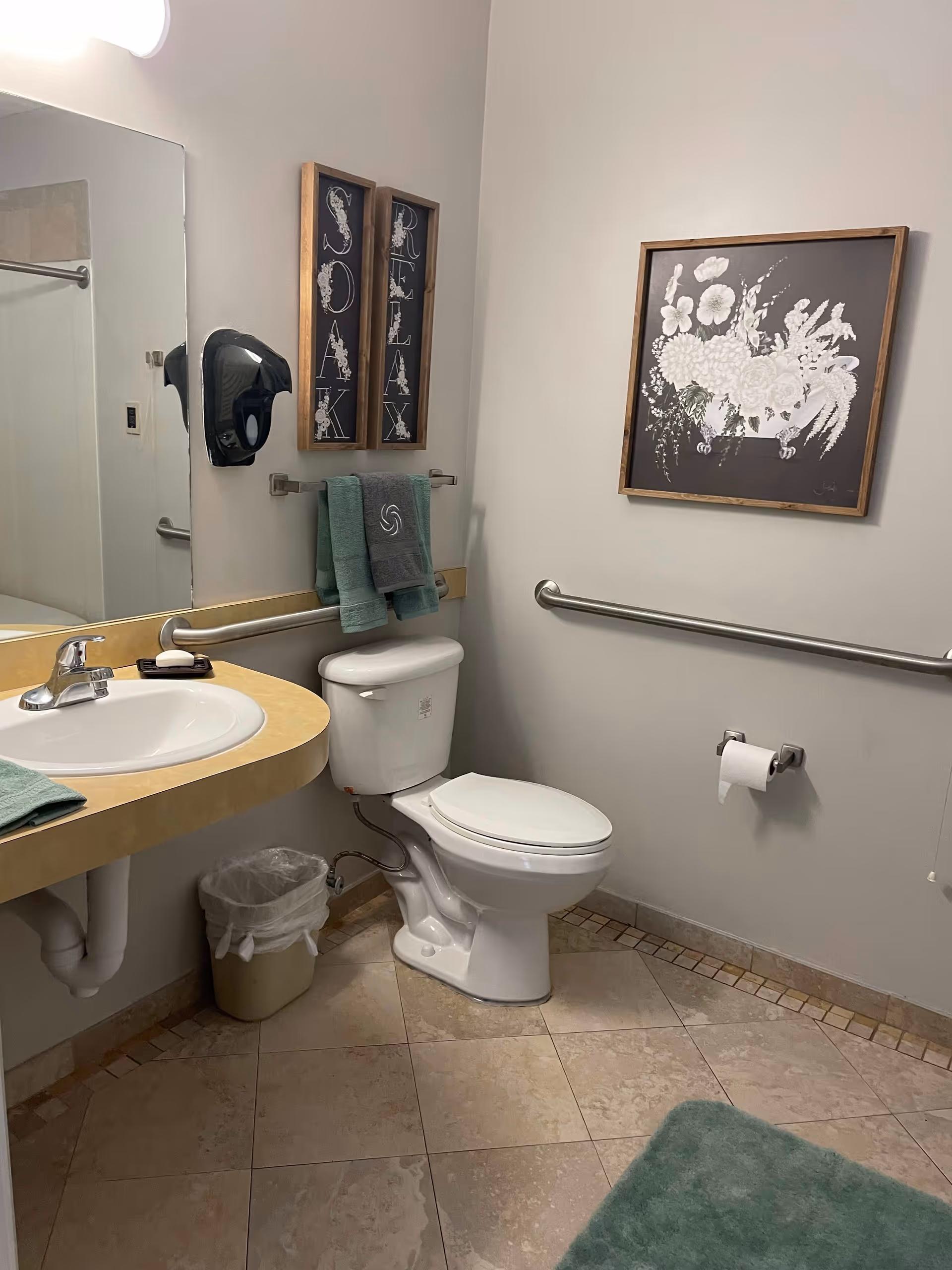 A clean bathroom with a white toilet and a beige countertop sink. There are two green towels hanging on a rack above the toilet, and a gray towel with a logo. On the wall above the towels are two vertical framed signs that say 'SOAK' and 'RELAX'. A framed floral artwork is hung on the adjacent wall. The bathroom has beige tiled flooring, a green bath mat, and a trash can under the sink. There are grab bars installed on the walls near the toilet and sink.
