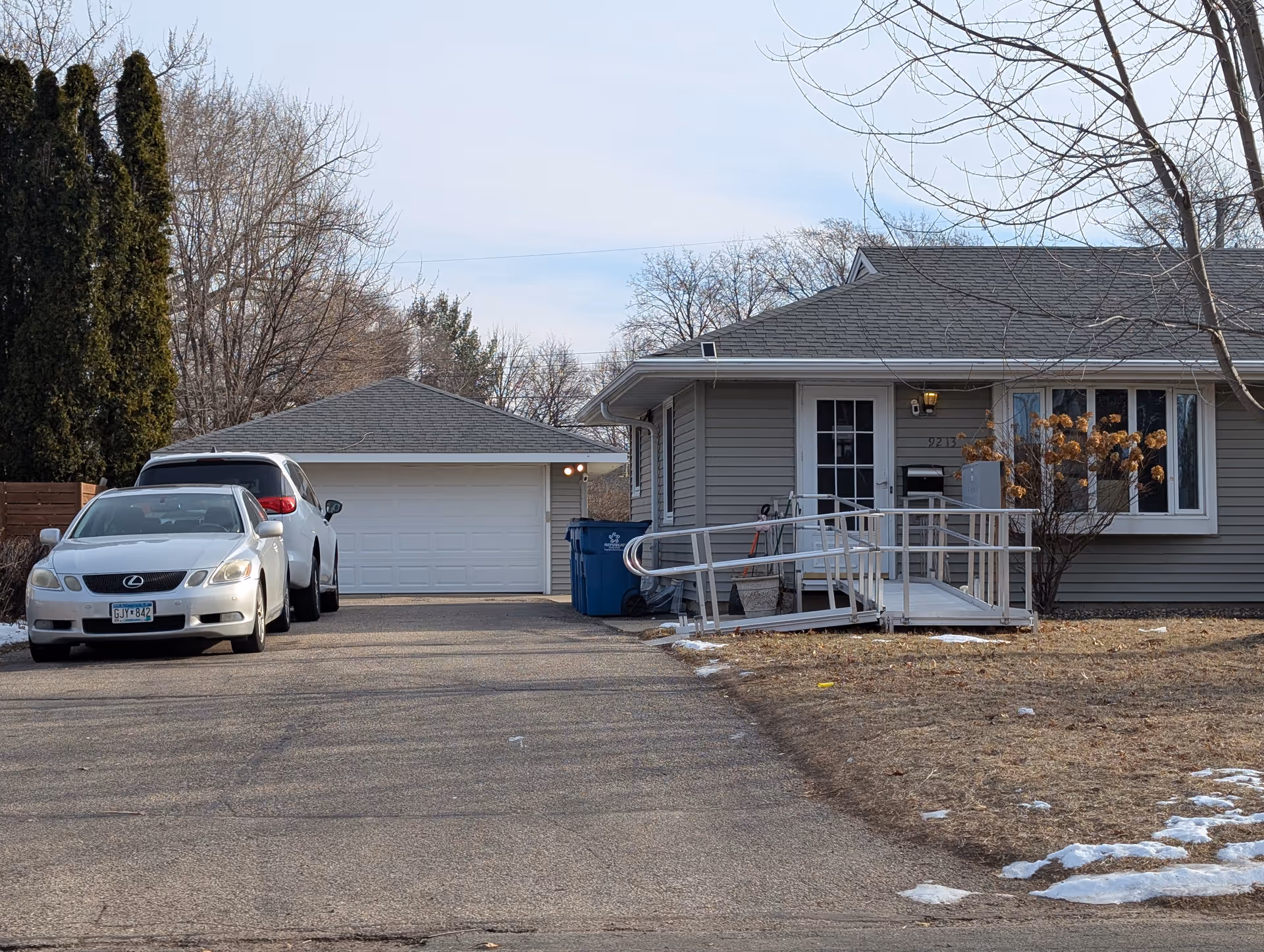 Single-story house exterior with a driveway, two parked cars, a detached garage, and a wheelchair ramp to the front door.