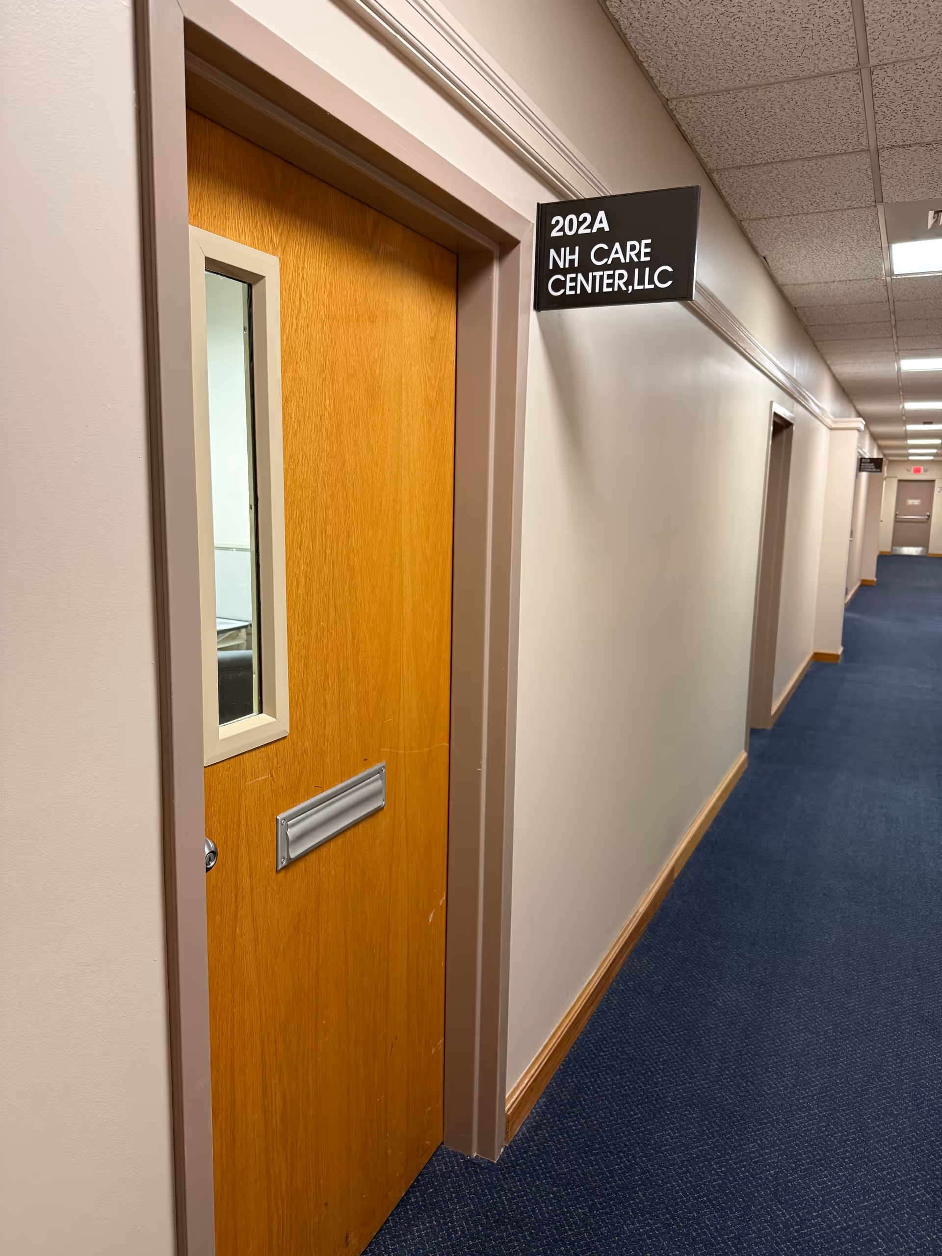 Interior hallway of NH Care Center with a wooden door labeled 202A along a blue carpet.