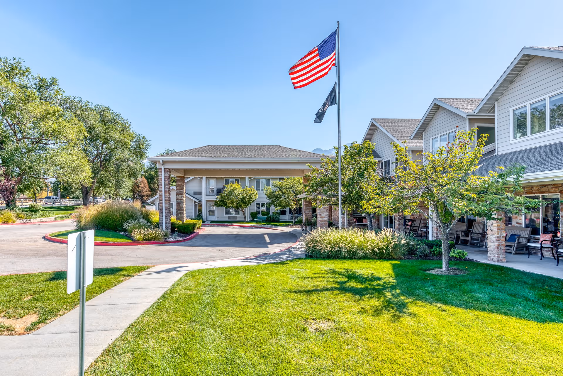 Exterior view of Solstice Senior Living at Sandy showing a well-maintained lawn, trees, a sidewalk, and a covered entrance with two flagpoles flying the American flag and another flag under a clear blue sky.