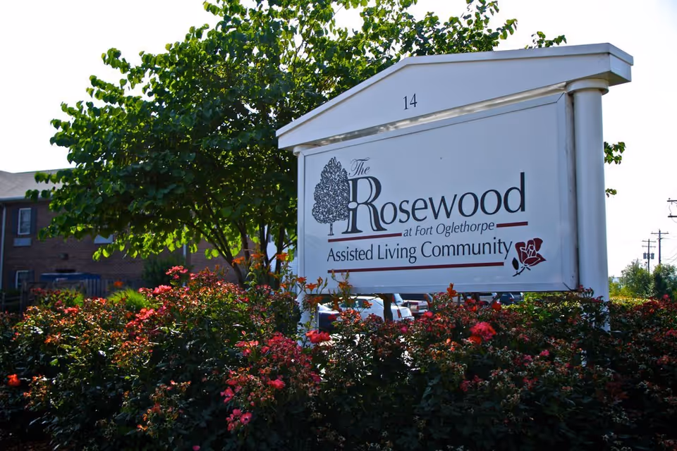 A white signboard reading The Rosewood at Fort Oglethorpe, surrounded by blooming bushes.