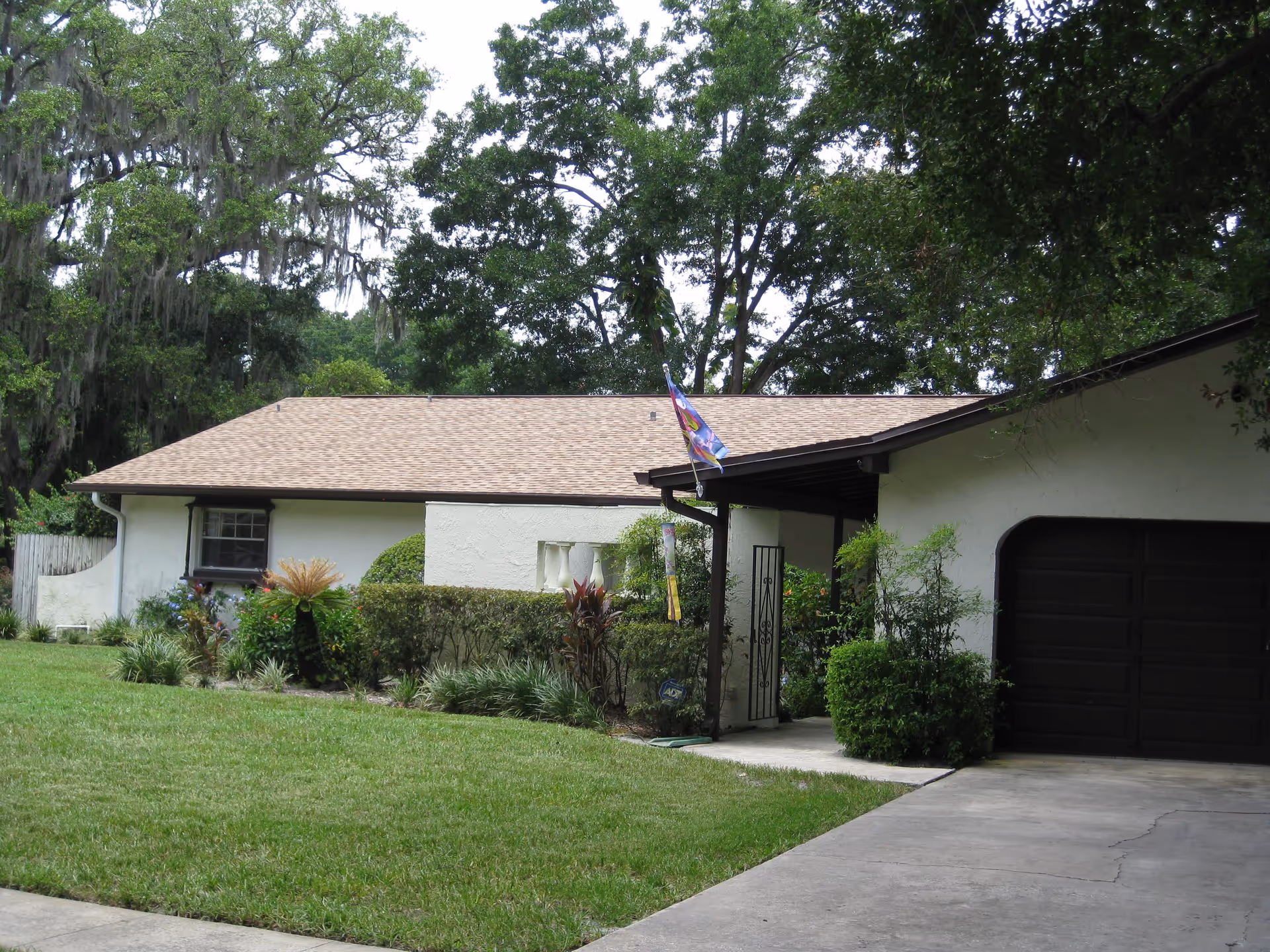 Front exterior of a single-story home with a garage, driveway, lawn, landscaping, and a flag beneath large trees.