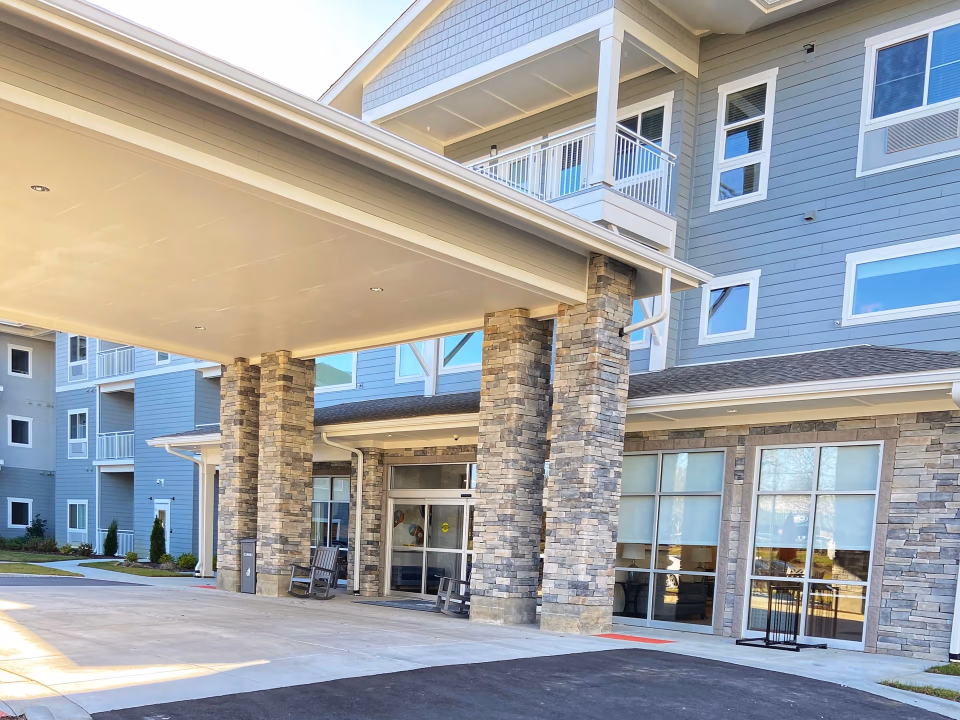 Entrance of a modern retirement community building with stone pillars supporting a covered drop-off area. The building has multiple floors with blue siding and large windows. There are rocking chairs near the entrance and a paved driveway in front.