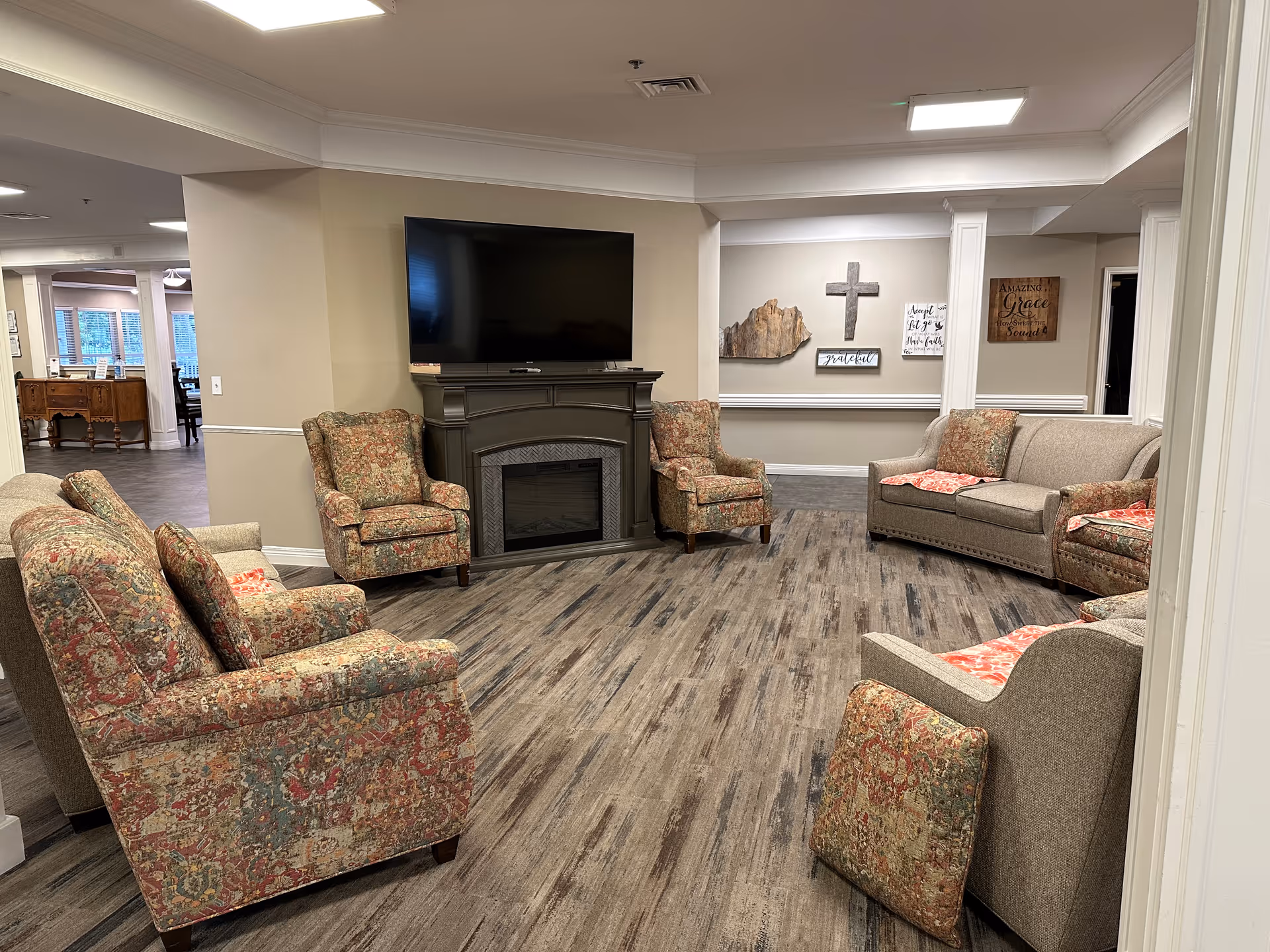 A cozy living room area in a senior living facility with patterned armchairs and beige sofas arranged around a dark fireplace with a mounted flat-screen TV above it. The room has wood-look flooring and neutral-colored walls decorated with inspirational wall art including a cross. The space is well-lit with ceiling lights and has an open view into an adjacent room with large windows.