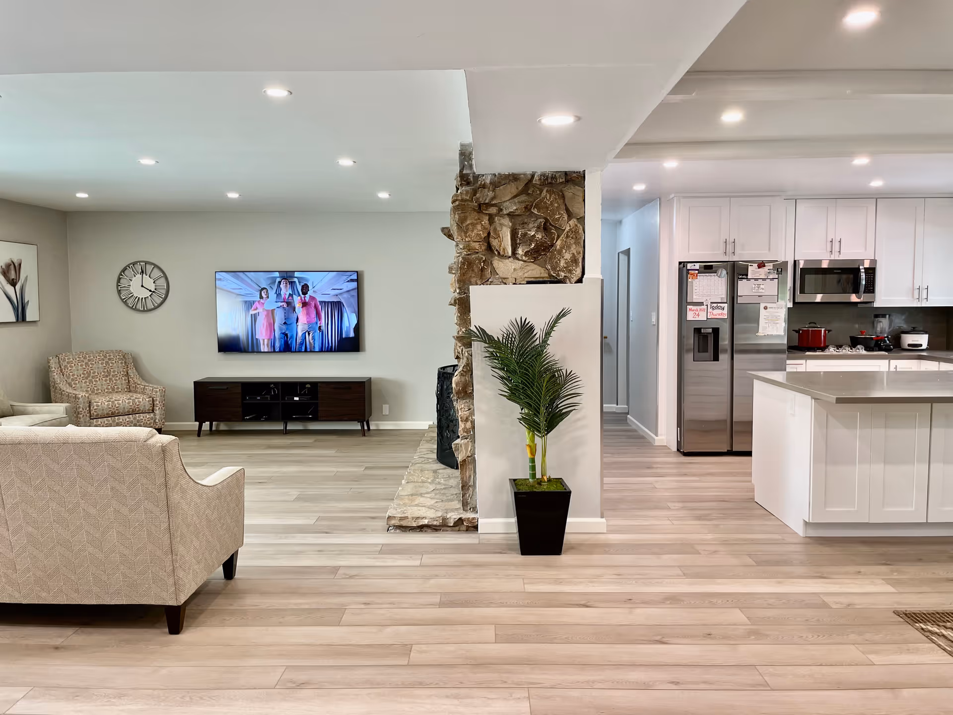 Bright open-plan living area with seating, TV, stone column, plant, and a nearby white kitchen.