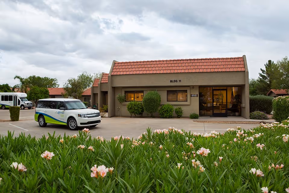 Exterior view of a single-story building labeled BLDG 11 with a red tiled roof and beige walls. There is a parking lot in front with a white and green vehicle parked. The building has a glass door entrance with a sign that reads 'Office'. Green bushes and flowering plants are visible in the foreground and around the building under a cloudy sky.