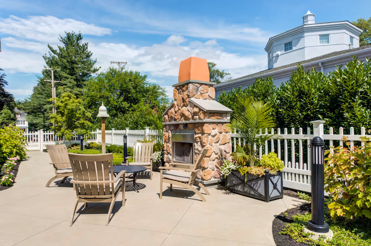 Sunny outdoor patio with a stone fireplace, Adirondack-style chairs around a small table, and a white picket fence with landscaping.