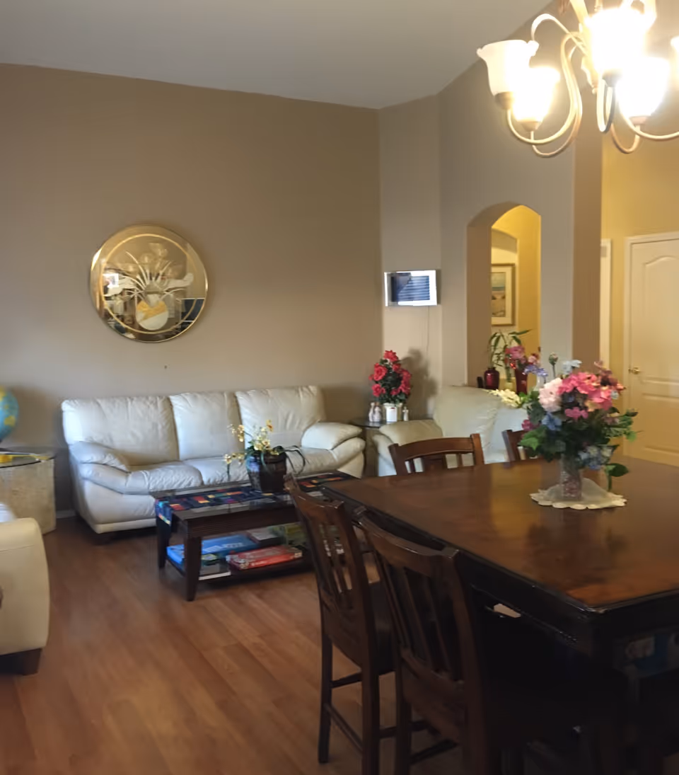 A spacious living room and dining area with white leather sofas, a wooden table, and flowers.