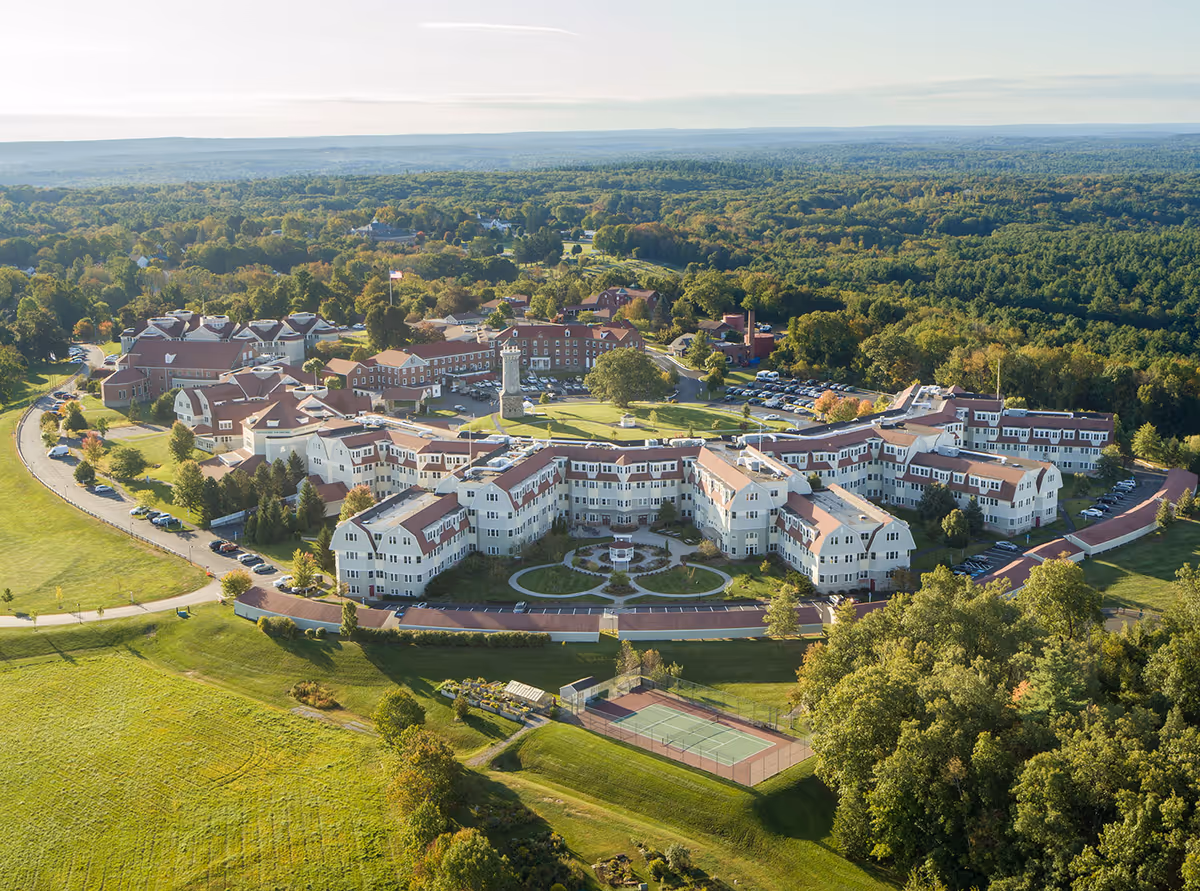 Aerial view of The Overlook senior living facility surrounded by greenery and trees, featuring multiple connected buildings with red roofs, a central courtyard with pathways and a gazebo, parking areas, and a tennis court in the foreground.