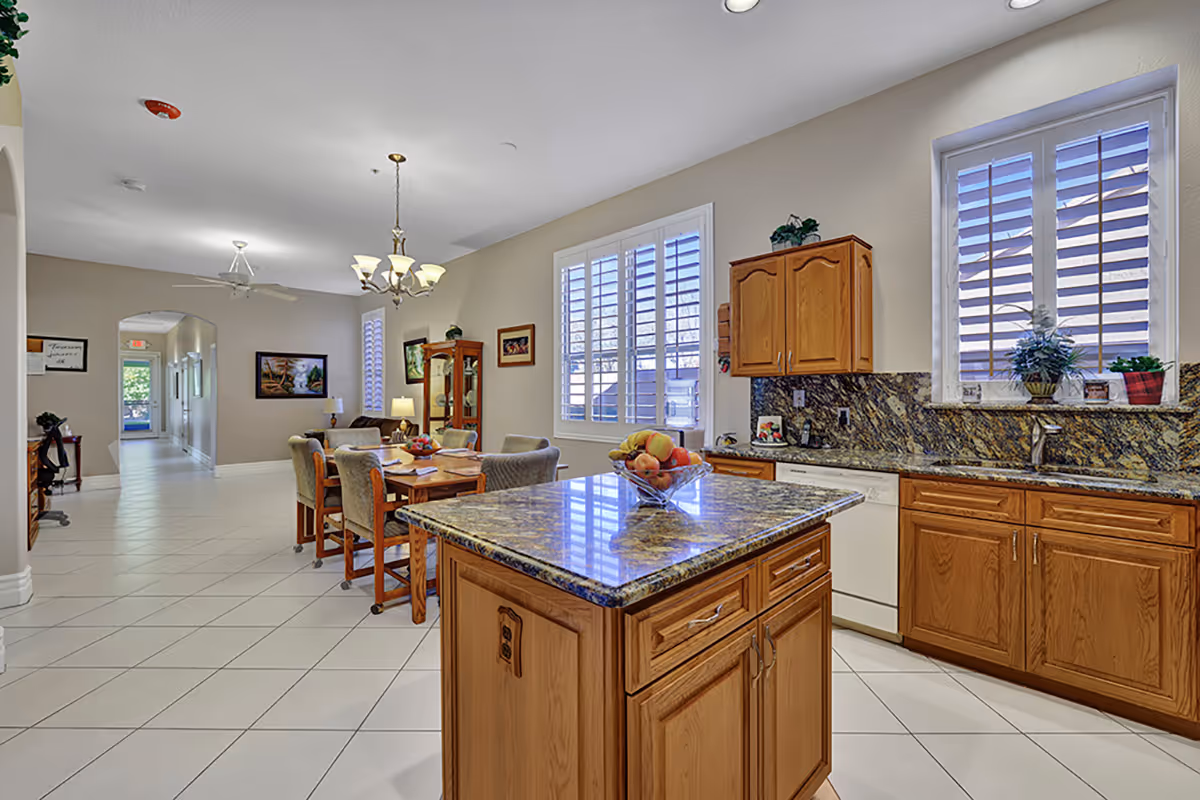 Spacious assisted living kitchen with granite island, wood cabinets, dining area, and large windows.