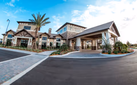 Front entrance of a large senior living clubhouse with a covered porte-cochere, palm trees, and a paved circular driveway under a blue sky.