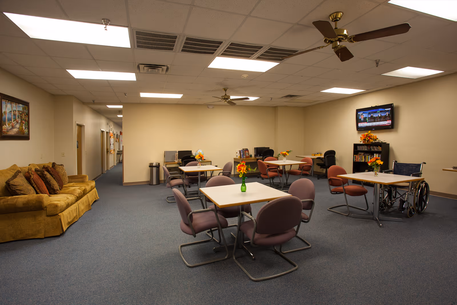 A spacious senior center lounge with round tables, purple chairs, a sofa, and a wall-mounted TV.