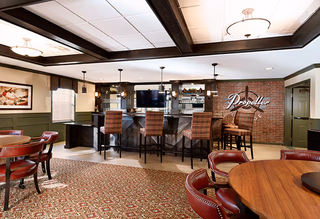 Interior view of a senior living facility lounge area featuring a bar with four high chairs upholstered in plaid fabric, a brick wall with a 'Propeller' sign, round wooden tables with red leather chairs, and a patterned carpet. The ceiling has recessed lighting and wooden beams.