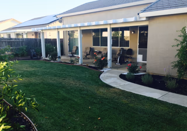 Outdoor view of a senior care facility showing a well-maintained lawn, a curved concrete pathway, and a covered patio area with chairs and exercise equipment. The building has beige walls and a roof with solar panels.