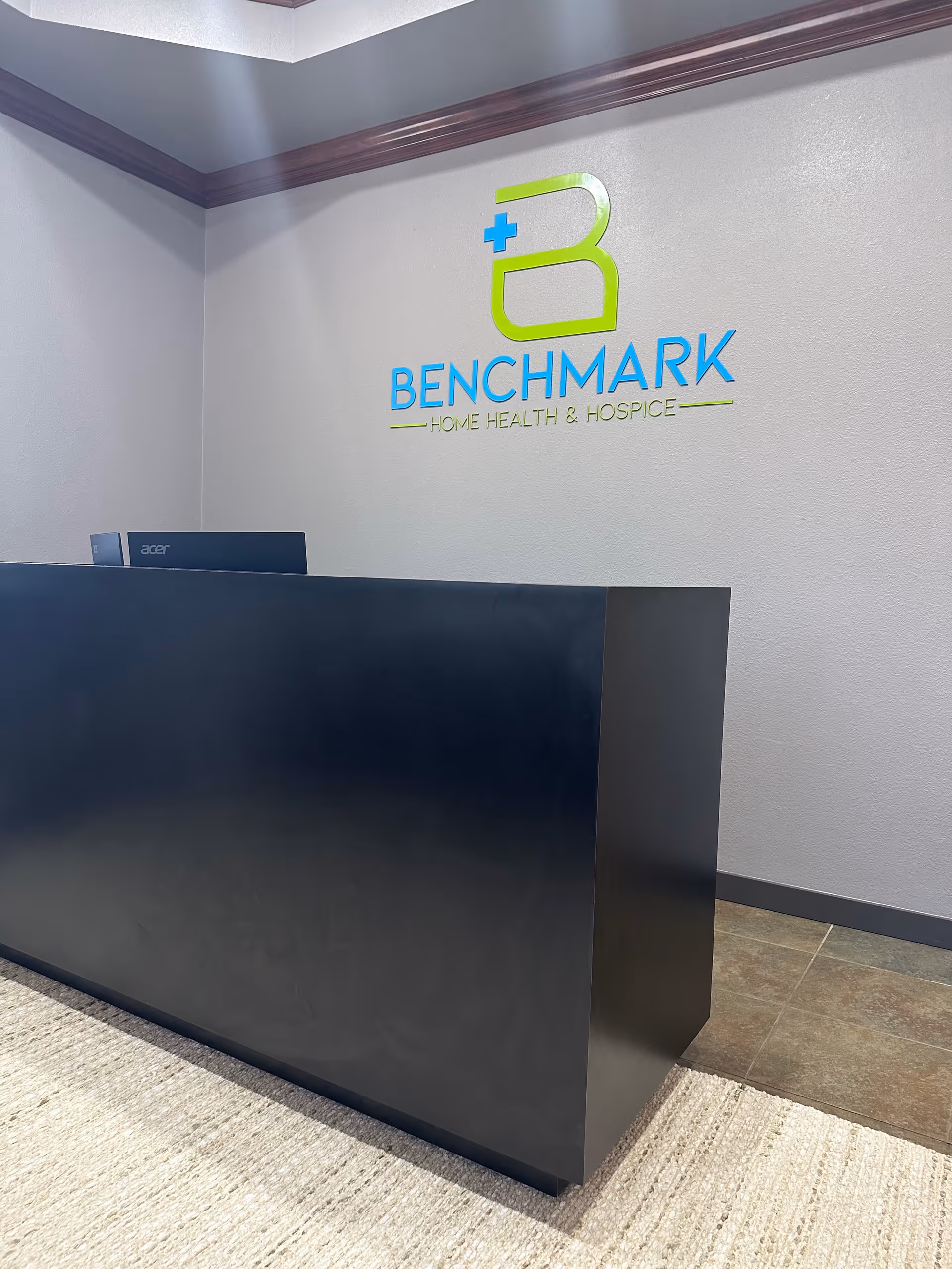 Lobby reception area with a dark curved desk and Benchmark Home Health & Hospice logo