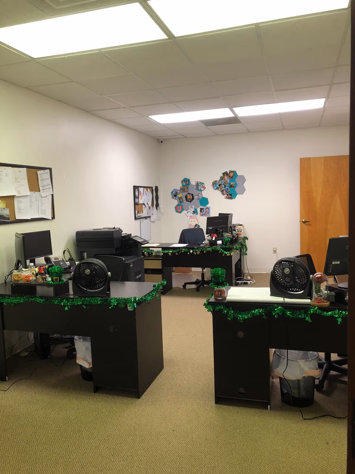 Office workspace with desks, computers, printers, and festive green garland decorations.