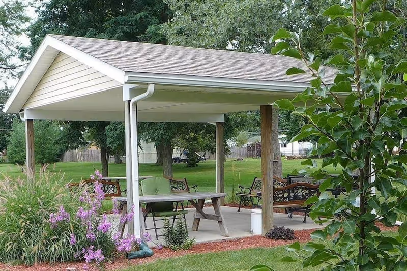 Outdoor covered pavilion with a picnic table and several benches surrounded by green grass, trees, and flowering plants in a garden setting.