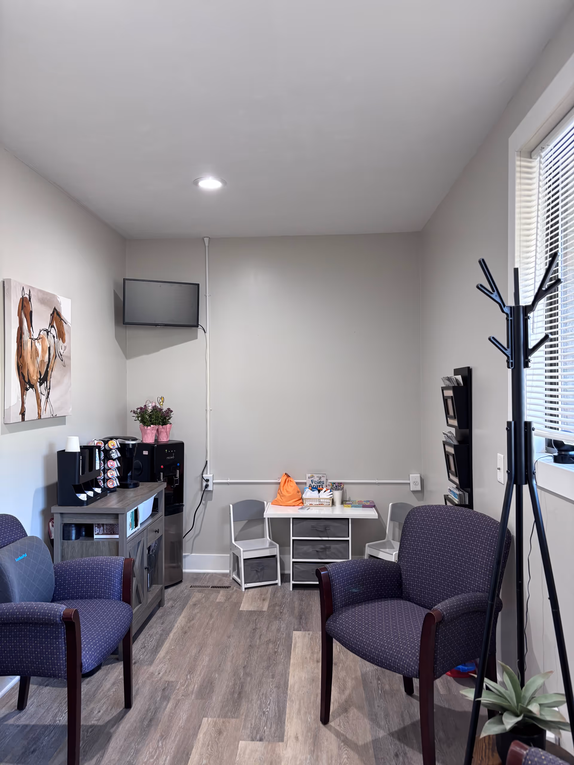 Small waiting lounge with two purple chairs, coffee machine, magazines, and a coat rack.