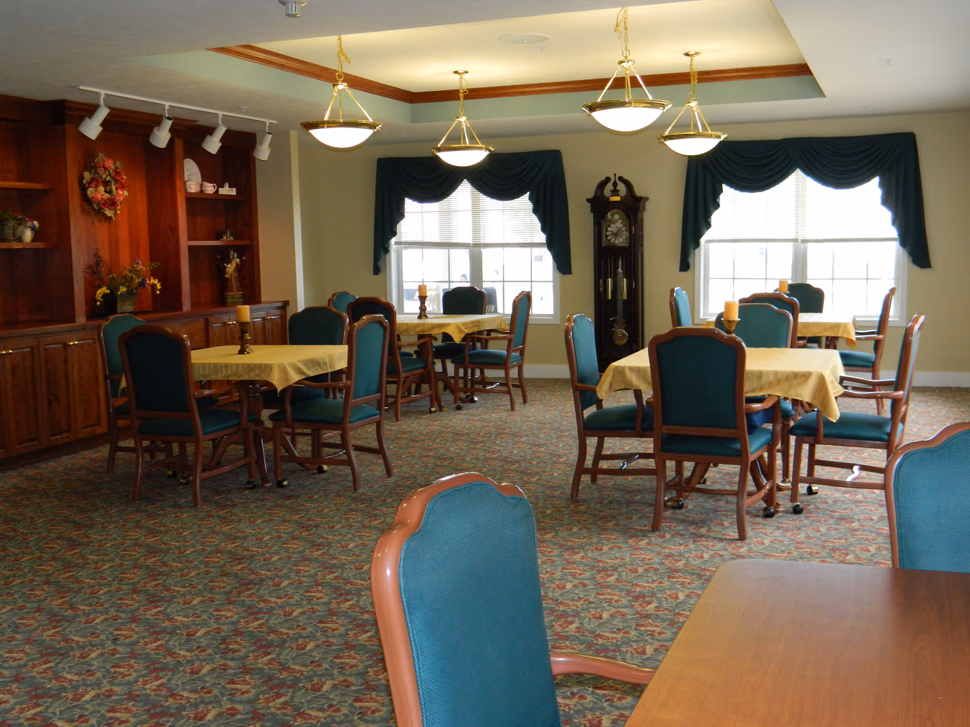 Spacious dining room with round tables, teal chairs, and decorative curtains in a senior facility.