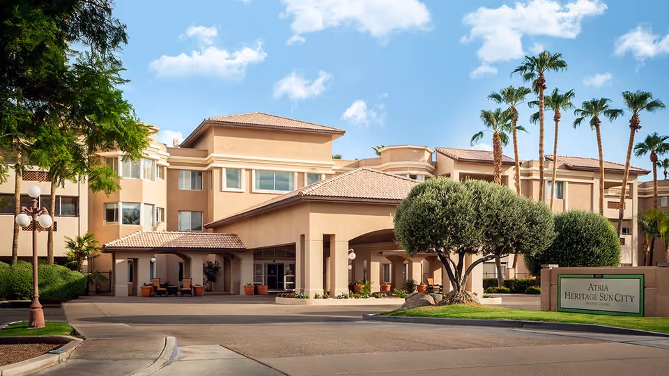 Exterior view of a senior living facility building with beige walls and a tiled roof under a blue sky with some clouds. The entrance has a covered driveway with potted plants and trimmed trees around the property. A sign in front reads 'ATRIA HERITAGE SUN CITY SENIOR LIVING'.