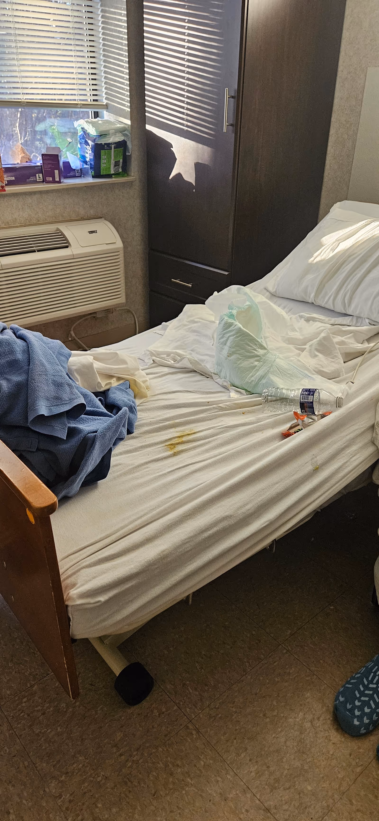Unmade bed in a small resident room with window blinds and wardrobe.