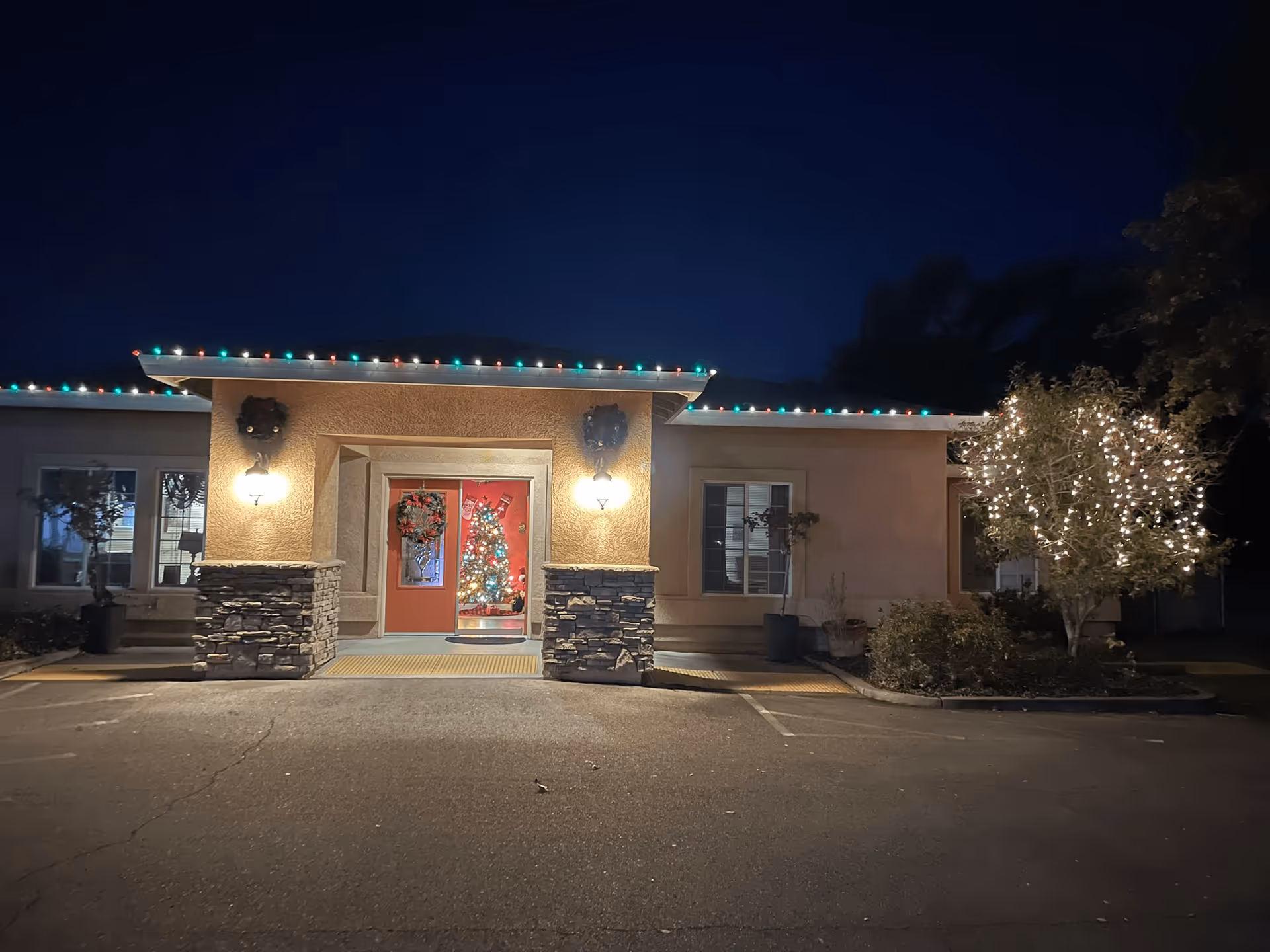 Night view of the front entrance of a building decorated with Christmas lights. The roofline is adorned with red, green, and white lights, and two wreaths hang on either side of the entrance. A Christmas tree with colorful lights is visible through the glass door. A tree on the right side of the entrance is wrapped in white string lights. The building has stone pillars flanking the entrance and windows on either side.