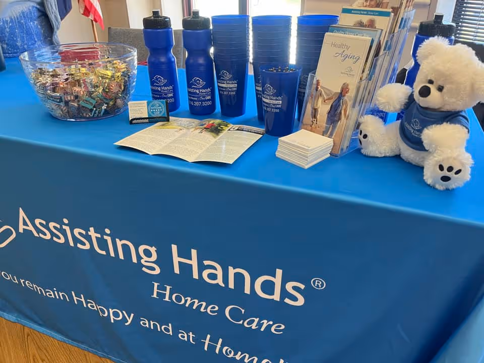 Promotional table with Assisting Hands Home Care branding, brochures, water bottles, and a teddy bear.