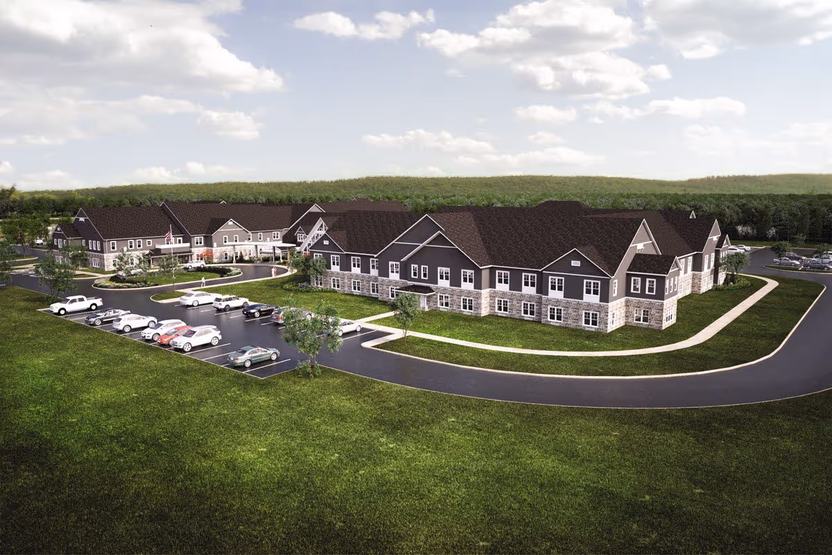 Aerial view of a large senior living facility building with dark roofs and light-colored walls, surrounded by green lawns and parking lots with several cars. The sky is partly cloudy.