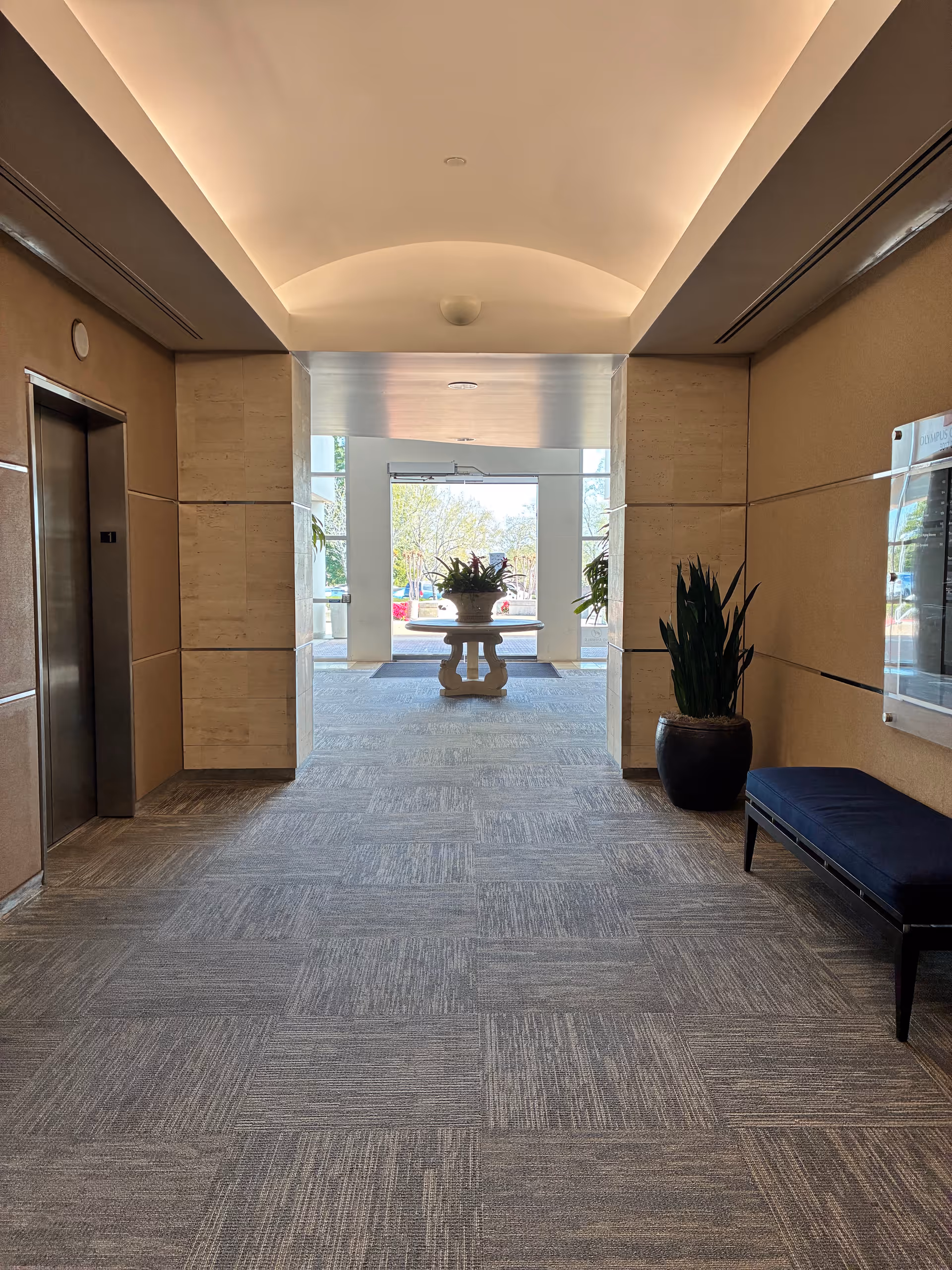 Interior hallway of a senior living facility with elevator, bench, and glass doors.